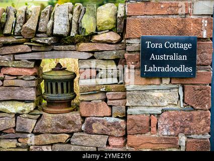 Ein schwarzes Schild mit weißem Text ist auf einer rustikalen Steinmauer montiert, auf dem der Name „Tora Cottage“ und die Hunderasse „Australian Labradoodles“ stehen. Die Stockfoto