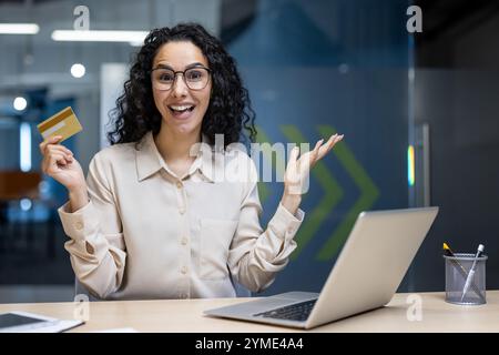 Hispanische Geschäftsfrau im Büro mit Kreditkarte, lächelnd mit Laptop. Steht für Online-Shopping, digitales Banking oder finanziellen Erfolg im professionellen Umfeld. Selbstbewusster, fröhlicher Ausdruck. Stockfoto