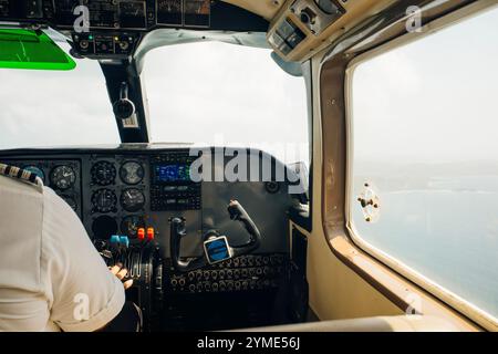 Nahaufnahme eines Mannes in einem Cockpit, der ein kleines viersitziges Flugzeug steuert und Knöpfe auf dem Bedienfeld drückt, mit Hügeln und Wiesen in der Ferne. Hoher ph-Wert Stockfoto