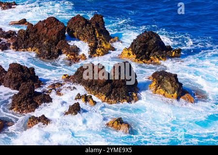 Pulsierende Küstenszene mit zerklüfteten Felsen, die aus dem Meer ragen. Wellen krachen gegen die Felsen und erzeugen weißen Schaum und Spritzer, der sich im Kontrast zum t befindet Stockfoto