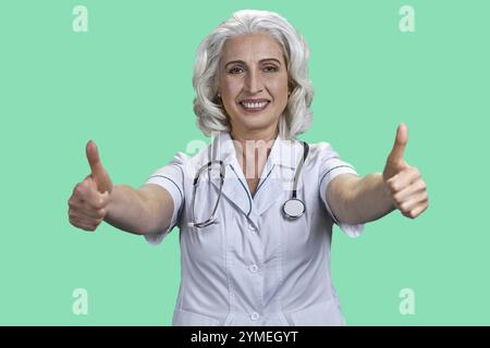 Schöne glückliche Ärztin, die mit beiden Händen das Daumenschild nach oben gibt. Weibliche Ärztin, die Daumen auf farbigem Hintergrund nach oben drückt Stockfoto