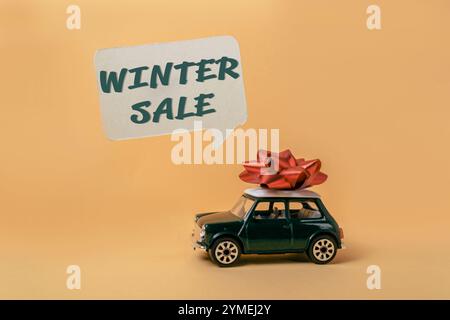 Das Spielzeugauto mit einer roten Schleife darauf sitzt auf einem gelben Hintergrund mit den Worten. Winterverkauf in einer Sprechblase geschrieben Stockfoto