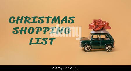 Grünes Spielzeugauto mit roter Schleife darüber. Das Auto ist auf gelbem Hintergrund mit den Worten Christmas Shopping List darunter Stockfoto