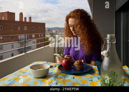 Junge Frau, die ihr Smartphone benutzt, während sie auf dem Balkon mit Blick auf die Stadt frühstückt Stockfoto