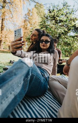 Junge Freunde, die einen sonnigen Tag im Freien genießen und gemeinsam Selfies auf einer Picknickdecke mit üppigen grünen Bäumen und Herbstlaub im Hintergrund machen Stockfoto