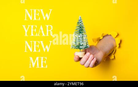 Hand hält einen kleinen Weihnachtsbaum vor einem gelben Hintergrund mit den Worten New Year New Me unten Stockfoto