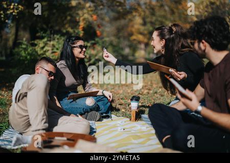 Gruppe von Freunden, die ein sonniges Picknick im Freien in einem Park genießen, plaudern und sich an einem wunderschönen Herbsttag entspannen. Sie sitzen auf einer Decke und Stockfoto