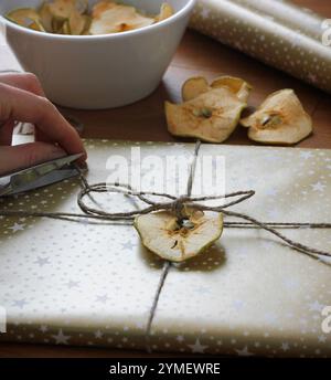 Nahaufnahme von weiblichen Händen, die Weihnachtsgeschenk umhüllen. Weihnachtsgeschenk in goldenem Geschenkpapier, verziert mit getrocknetem Apfel, getrockneten Äpfeln in einer Schüssel Stockfoto