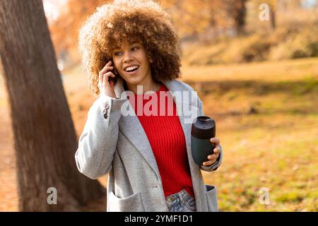 Helles Herbstlaub umgibt eine junge Frau, die lächelt und sich mit einem warmen Getränk in der Hand unterhält. Sie verkörpert einen freudigen Moment Stockfoto