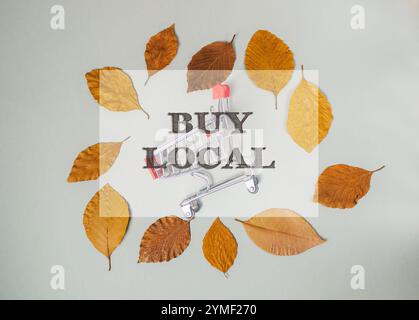 Der Einkaufswagen ist von Blättern umgeben und die Worte Buy Local stehen darüber. Konzept der Gemeinschaft und Unterstützung lokaler Unternehmen Stockfoto