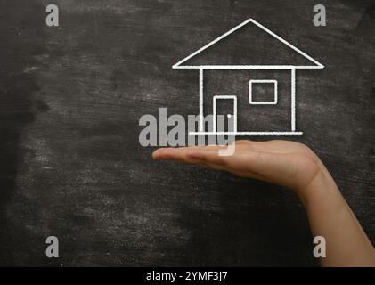 Hand hält eine Kreidezeichnung eines Hauses. Die Zeichnung steht auf einer Tafel. Das Haus ist in weißer Kreide gezeichnet Stockfoto