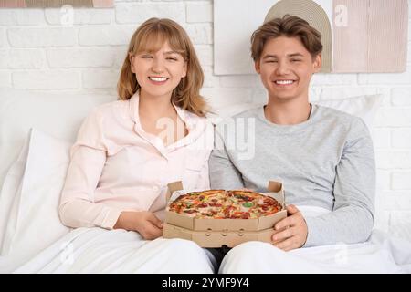 Wunderschönes junges Paar mit Pappkarton mit leckerer Pizza und auf dem Bett im Schlafzimmer Stockfoto