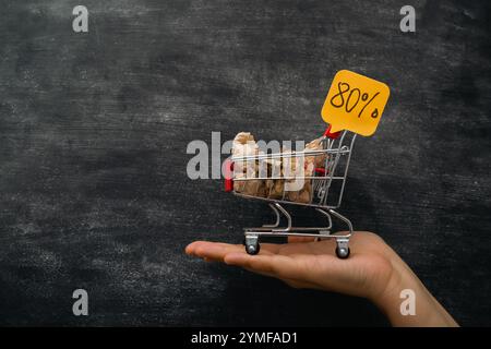 Hand hält einen Einkaufswagen mit einem Schild, auf dem 80 steht. Der Warenkorb ist mit Artikeln gefüllt, und die Person hält ihn hoch, um den Rabatt anzuzeigen Stockfoto