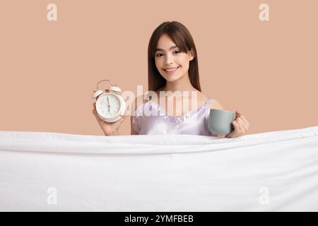 Schöne junge Frau mit Wecker, Tasse Kaffee und Laken auf beigefarbenem Hintergrund Stockfoto