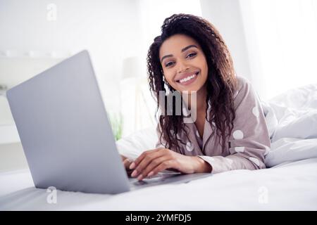 Junge Frau mit Afro-Haar im Pyjama mit Punktmuster lächelt hell, während sie im gemütlichen Schlafzimmer einen Laptop benutzt Stockfoto