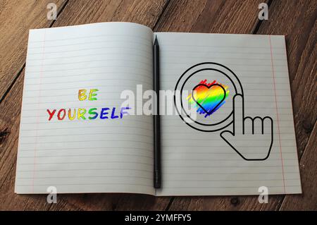 Notizbuch mit einem Regenbogenherz und den Worten „Be Yourself“ darauf. Auf der Seite wird ein schwarzer Stift platziert Stockfoto