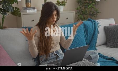 Eine junge brünette Frau sitzt in einem gemütlichen Wohnzimmer auf einer Couch mit Laptop und drückt Verwirrung oder Frustration aus, während sie von zu Hause aus arbeitet. Stockfoto