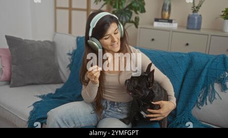 Eine junge hispanische Frau im Wohnzimmer hört Musik mit Kopfhörern, während sie ihre französische Bulldogge auf der Couch kuschelt. Stockfoto