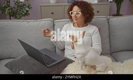 Eine reife Frau meditiert auf einer Couch mit Laptop und einem chihuahua in einem gemütlichen Wohnzimmer. Stockfoto