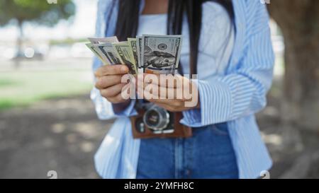 Eine junge hispanische Frau steht in einem Park und zählt uns Dollarscheine, während sie eine Kamera um den Hals hält und eine Szene im Freien zeigt. Stockfoto