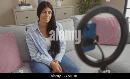 Eine junge, attraktive hispanische Frau, die sich in ihrem Wohnzimmer filmt, auf einer Couch sitzt, mit einem Smartphone mit Ringlicht ein gemütliches und persönliches Ambiente schafft Stockfoto