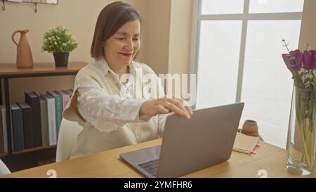 Eine Frau mittleren Alters benutzt zu Hause einen Laptop und lächelt, während sie potenziell mit jemandem interagiert, der in einer gemütlichen Einrichtung online ist. Stockfoto