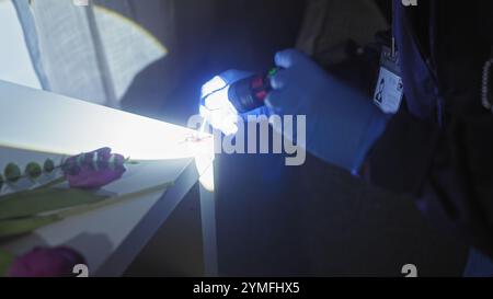 Ein forensischer Experte untersucht einen dunklen Tatort im Innenbereich mit einer Taschenlampe und Beweismitteln auf einem Tisch. Stockfoto