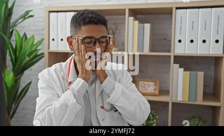 Gestresster afroamerikanischer Arzt mit Brille in einer Krankenhauspraxis mit Händen im Gesicht. Stockfoto