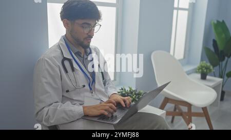 Gutaussehender arabischer Mann in Brille und Stethoskop mit Laptop im modernen Klinikbüro Stockfoto