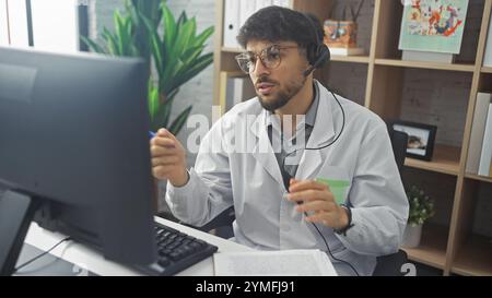 Ein junger bärtiger Mann in einem weißen Labormantel mit Headset konzentriert sich in einer modernen Klinik auf einen Computerbildschirm. Stockfoto