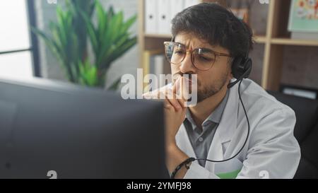 Ein nachdenklicher arabischer Mann mit Bart und Brille, der ein Headset und einen Labormantel in einer modernen Büroumgebung trägt, was auf eine medizinische Beratung hindeutet. Stockfoto