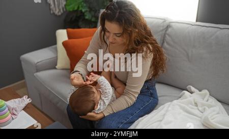 Mutter und Tochter im Wohnzimmer teilen sich liebevolle Momente auf dem Sofa, Frau, die Ihr Baby mit Zärtlichkeit in gemütlicher Wohnatmosphäre mit warmer Einrichtung und weicher Note versorgt Stockfoto