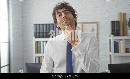 Junger Mann in Hemd und Krawatte steht selbstbewusst in einem modernen Büro mit Bücherregalen und großem Fenster mit Tageslicht. Stockfoto