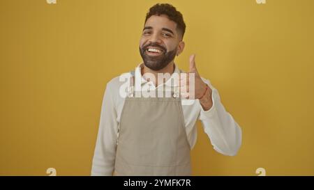 Lächelnder bärtiger Mann auf Schürze, der die Daumen gegen die gelbe Wand hält Stockfoto