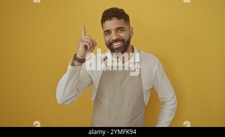 Lächelnder bärtiger hispanischer Mann in Schürze, der isoliert auf gelbem Hintergrund nach oben zeigt Stockfoto