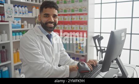 Ein lächelnder junger Mann mit Bart, der drinnen an einer Apotheke arbeitet, mit einem Computer. Stockfoto