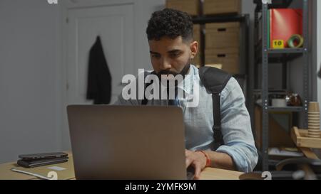 Konzentrierter hispanischer Mann im Büro, der an einem Laptop arbeitet, mit Regalen, professioneller Kleidung und potenzieller Detektivatmosphäre Stockfoto