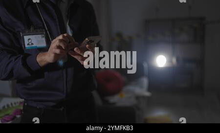 Detective untersucht Beweise mit dem Smartphone im Scheinwerferlicht an einem dunkel beleuchteten Tatort in Innenräumen Stockfoto