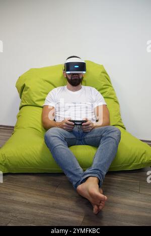 Mann mit VR-Headset, bequem auf grünem Sitzsack sitzend, während er mit dem Controller spielt Stockfoto
