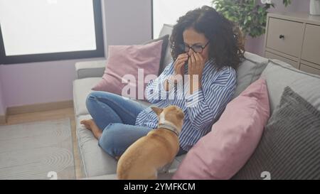 Eine hispanische Frau mittleren Alters sitzt drinnen mit ihrem chihuahua auf einer Couch und weint, während sie von Kissen umgeben ist, in einem gut beleuchteten Wohnzimmer. Stockfoto