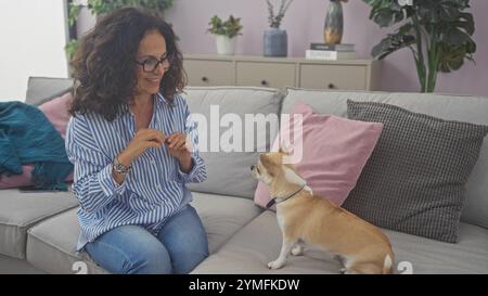 Eine Frau mittleren Alters interagiert mit ihrem chihuahua in einem gemütlichen, gut eingerichteten Wohnzimmer und schafft so eine gemütliche Atmosphäre. Stockfoto