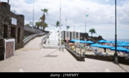 Unscharfer Blick auf einen Strand mit blauen Sonnenschirmen, Steinweg und verschwommenem Hintergrund des Ozeans und Palmen unter einem bewölkten Himmel. Stockfoto