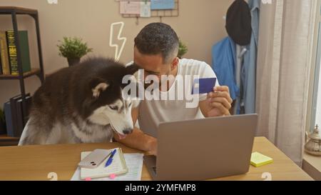 Ein fröhlicher junger hispanischer Mann mit Kreditkarte und Laptop sitzt drinnen neben seinem aufmerksamen Huskyhund und unterstreicht eine ungezwungene und gemütliche Atmosphäre. Stockfoto