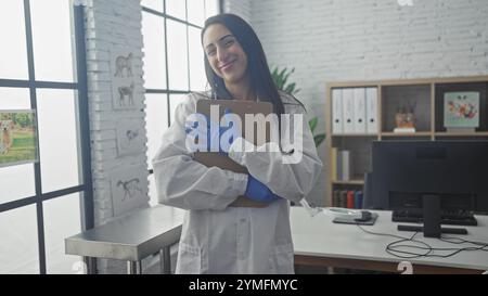 Eine lächelnde junge hispanic-Tierärztin umarmt ein Klemmbrett in einem hellen Klinikbüro Stockfoto