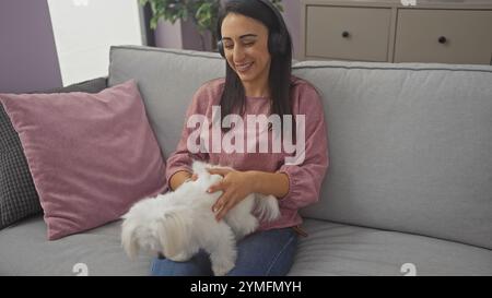 Hispanische Frau genießt Musik mit Kopfhörern und kuschelt einen maltesischen Hund auf einem Sofa in einem gemütlichen Wohnzimmer. Stockfoto