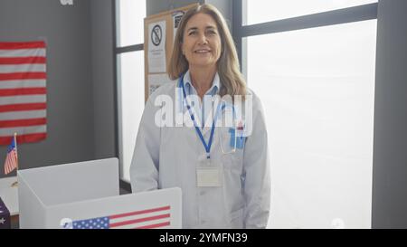 Ärztin mittleren Alters lächelt in einer amerikanischen Wahlstation mit Flagge und Wahlurne Stockfoto