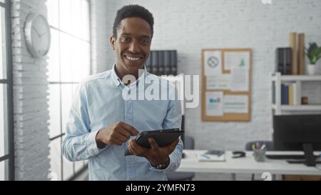 Junger Mann lächelt in hellen Büroumgebungen mit einem Tablet, das professionelle und moderne Arbeitsplatzumgebung mit afroamerikanischen Männern zeigt Stockfoto