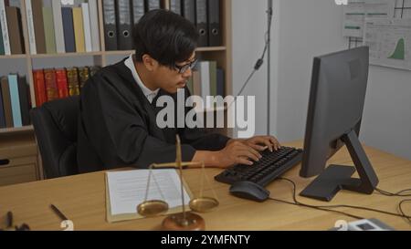 Junger Mann in richterlichem Gewand, der an seinem Schreibtisch in einem Büro mit juristischen Büchern und Waagen in einem professionellen Gerichtssaal arbeitet. Stockfoto