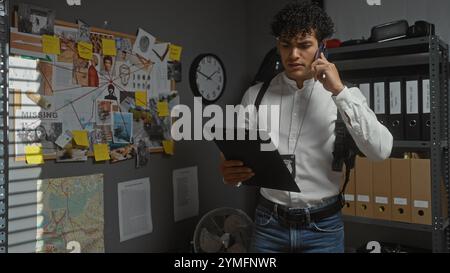 Hispanischer Mann, der am Telefon spricht, während er im Büro eines Detektivs eine Zwischenablage mit Beweismaterial, Uhr und Aktenschränken hält. Stockfoto