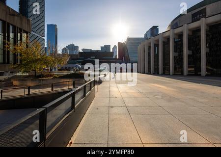 Das Bild zeigt die Place des Arts in Montreal bei tiefstehender Sonne. Der weite Platz aus geometrisch angeordneten Betonplatten wird von modernen Gebäuden und einer Skulptur aus reflektierenden Kugeln eingerahmt. Die warmen Sonnenstrahlen erzeugen ein weiches Licht- und Schattenspiel auf dem Boden, das eine warme, ruhige Atmosphäre vermittelt. Auf der rechten Seite befinden sich markante architektonische Säulen des Opernhauses, während links ein einzelner Baum in herbstlichen Farben zu sehen ist. Eine einzelne Person auf einem Roller fügt eine narrative Komponente hinzu und belebt die Szene s Stockfoto